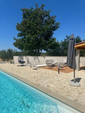 Ein Swimmingpool mit Liegestühlen und Sonnenschirm, umgeben von einer Steinterrasse und einem großen Baum.