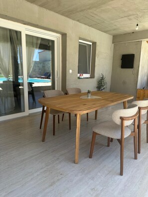 Ein geräumiger Raum mit einem großen Holztisch, umgeben von Stühlen, einer Glastür zum Poolbereich und einem an der Wand befestigten Fernseher.