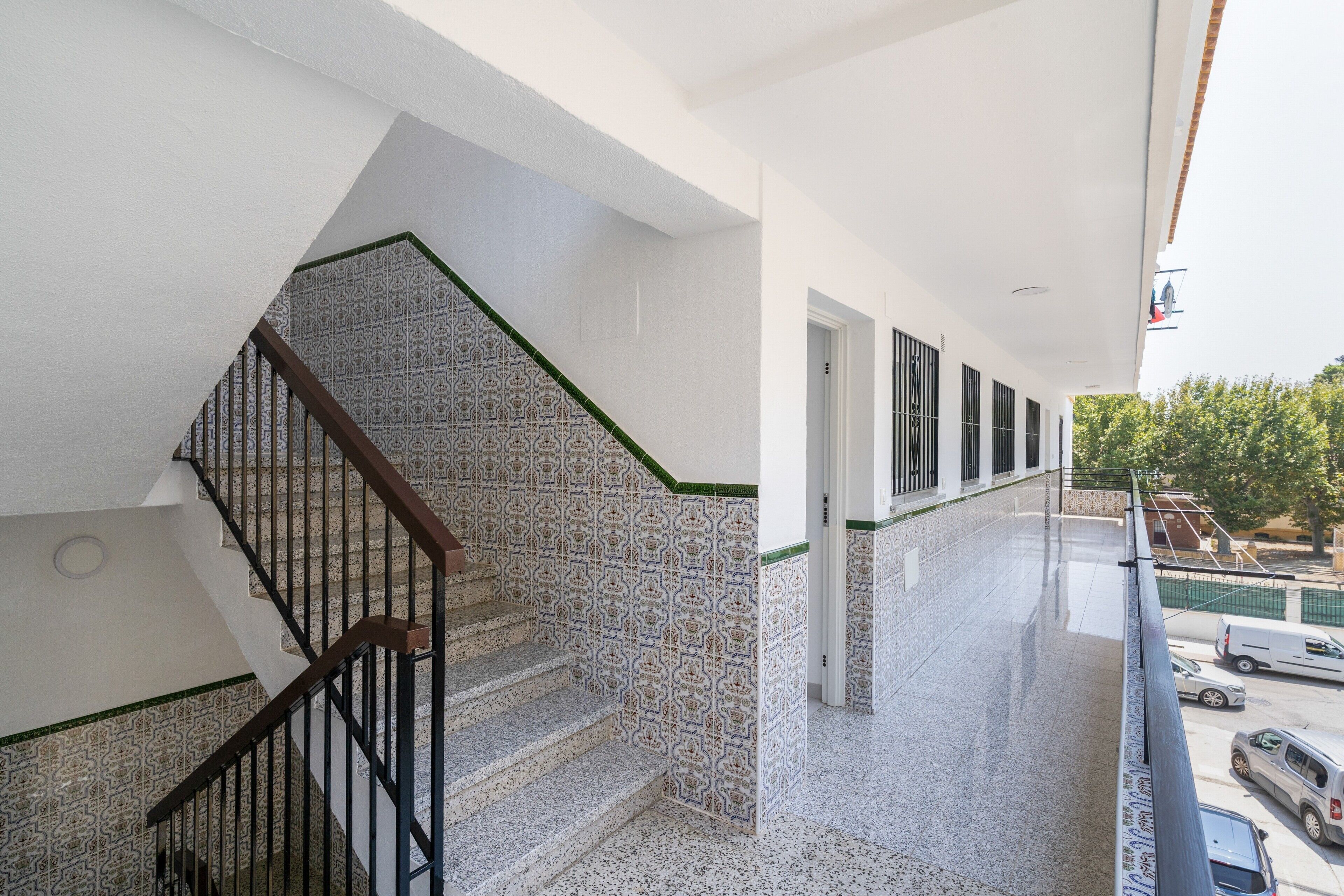 A staircase with patterned tiles, a balcony with a view of parked cars, and a building with a white exterior.