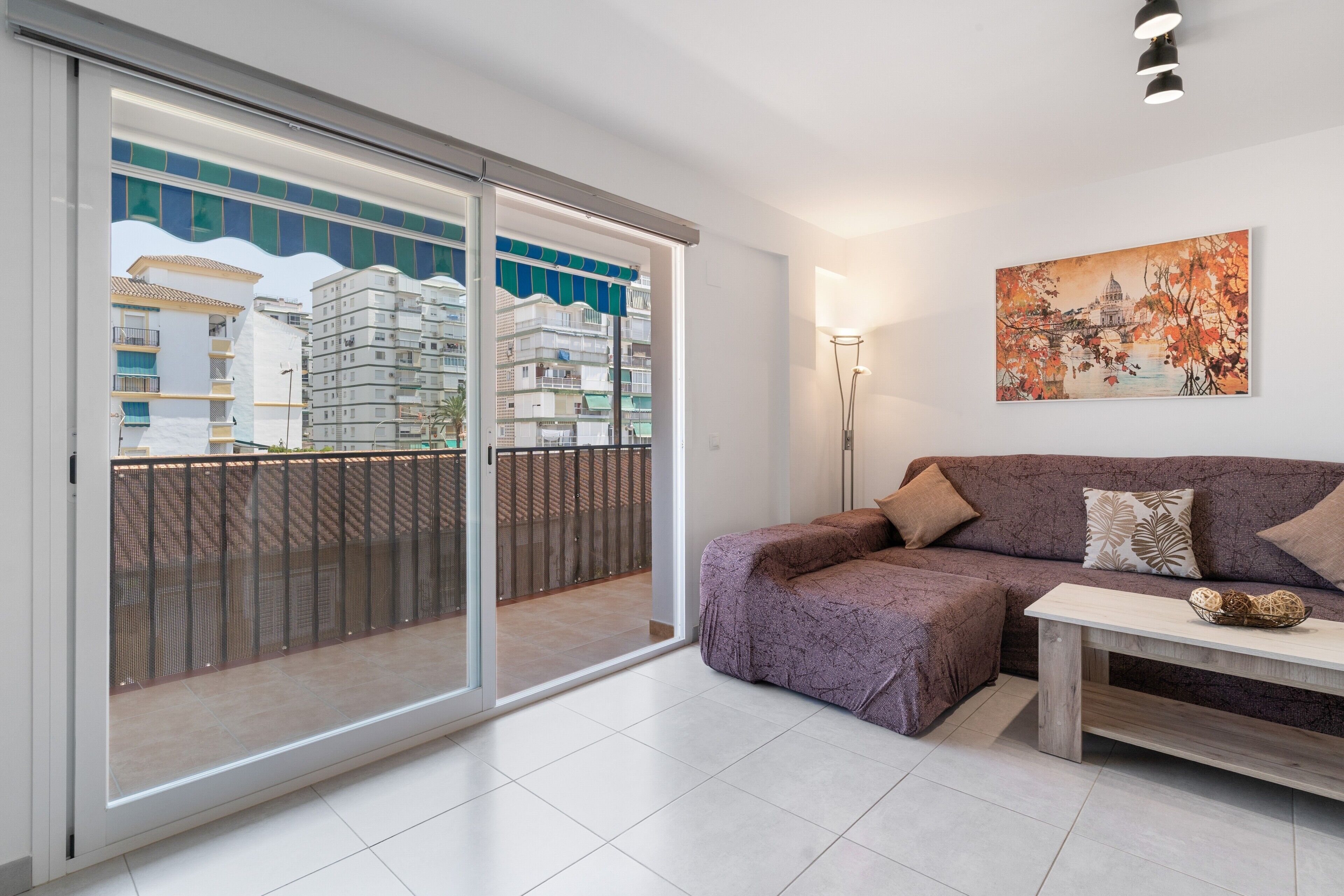 A modern living room with a sliding glass door leading to a balcony, a purple sofa set, a floor lamp, and a painting on the wall.