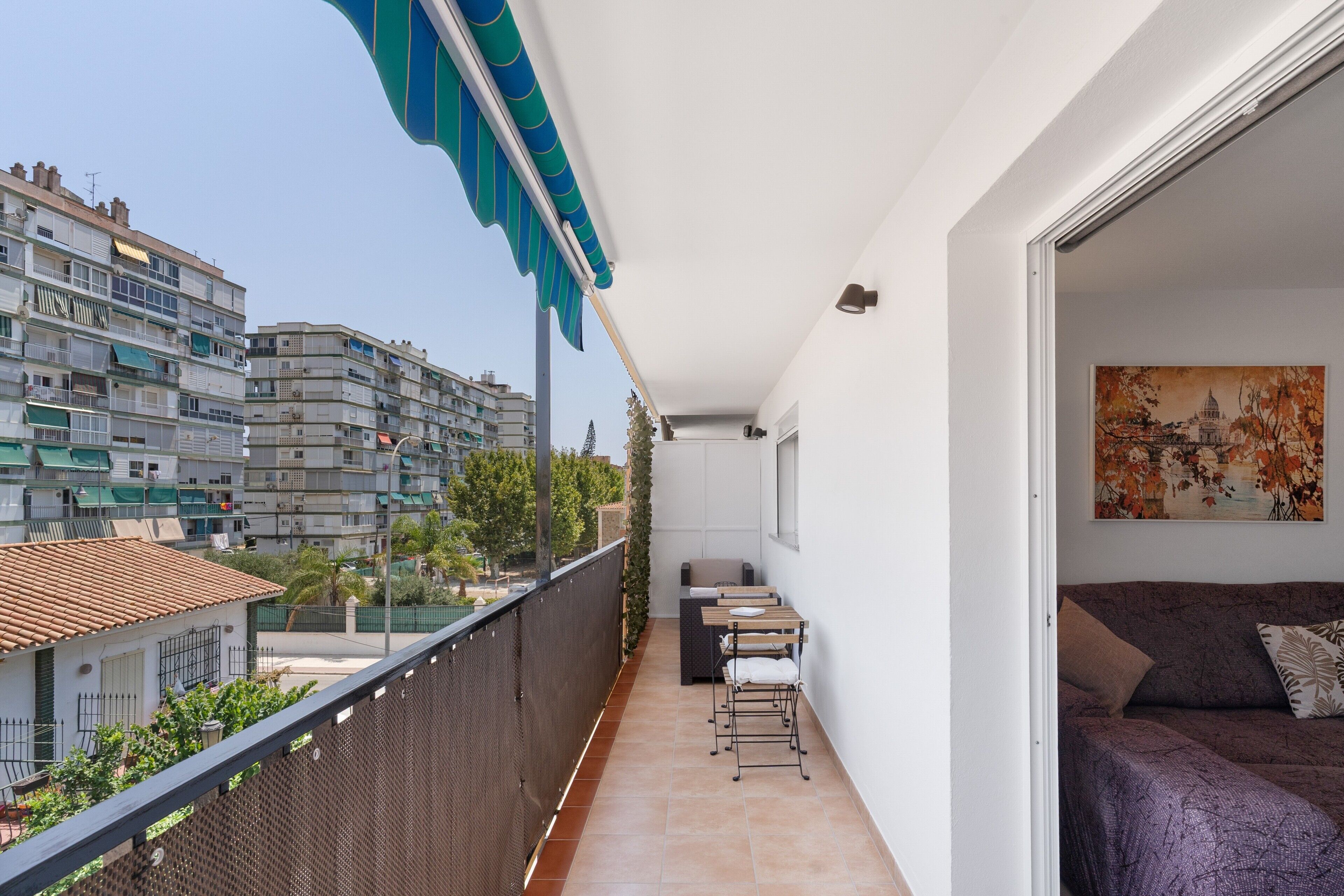 Un balcón con vistas a la ciudad, un pequeño espacio de descanso y una puerta que da a una habitación con una cama y un cuadro.