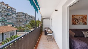 A balcony with a view of a cityscape, a small seating area, and a door leading to a room with a bed and a painting.