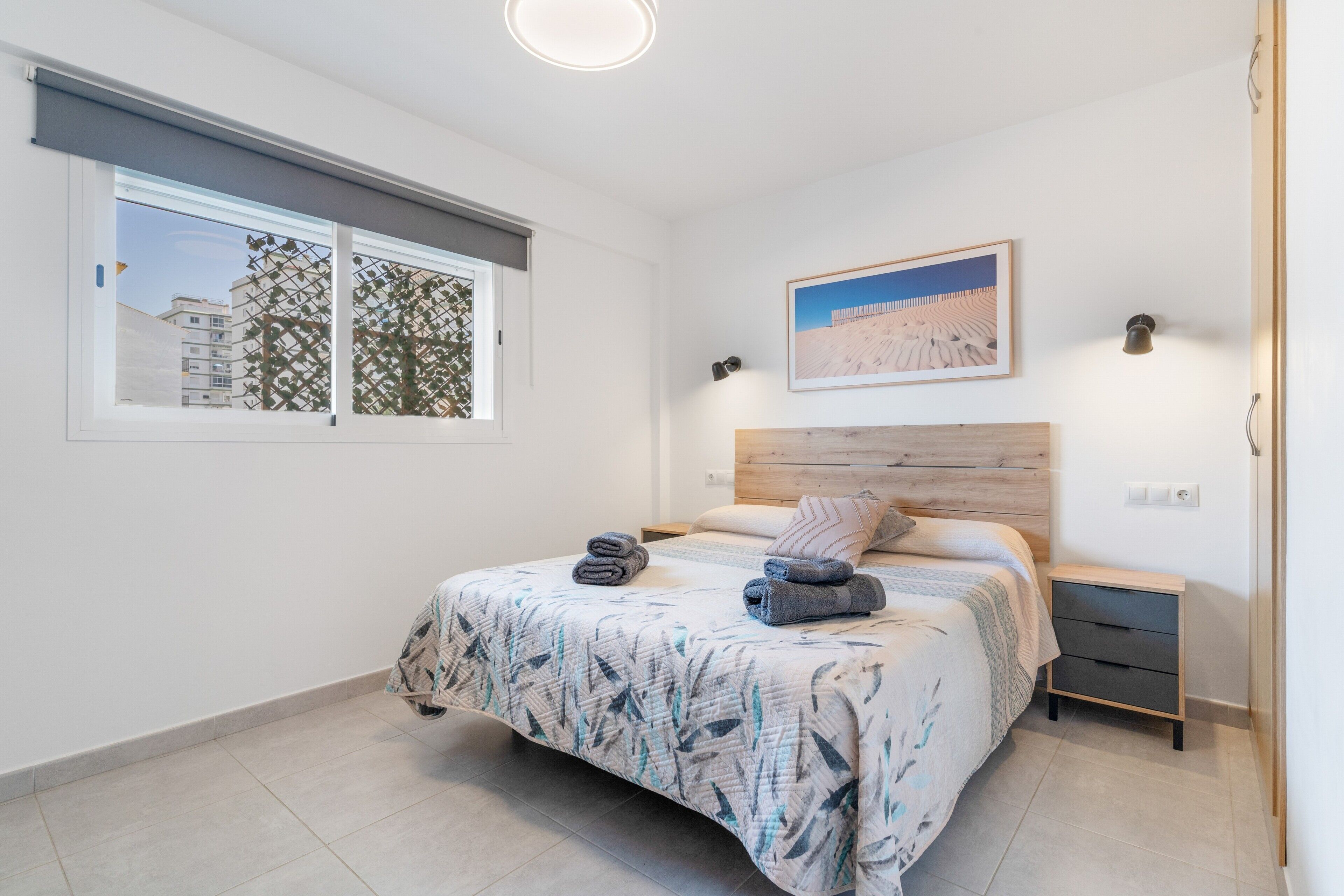 Un dormitorio con cama, ventana con vistas a edificios, un cuadro colgado en la pared y una mesita de noche.