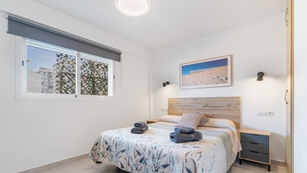 A bedroom with a bed, a window with a view of buildings, a wall-mounted picture, and a bedside table.