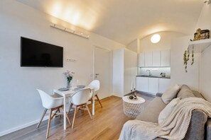 Una sala de estar moderna con un sofá, una mesa de comedor con sillas, un televisor montado en la pared y una zona de cocina con armarios y un fregadero.