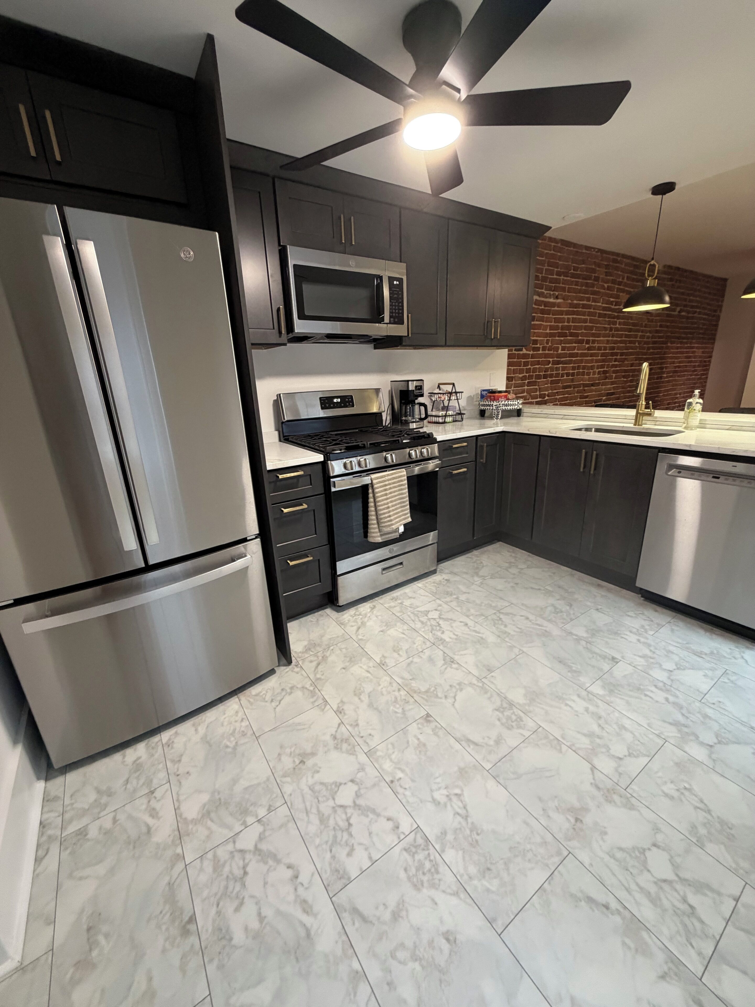 A modern kitchen with stainless steel appliances, a brick accent wall, and a ceiling fan.