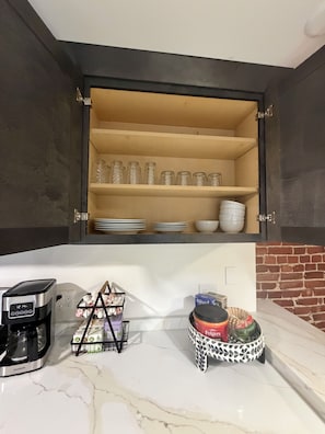 Une cuisine équipée d’une machine à café, d’un panier rempli d’ustensiles et d’une étagère où sont rangés des bocaux en verre et de la vaisselle.