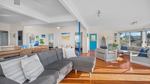 Ein modernes Wohnzimmer mit einem grauen Ecksofa, einem Essbereich mit Holztisch und Stühlen sowie einer hellen Küche mit weißen Schränken.