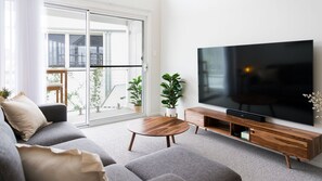 A modern living room with a large flat-screen TV, grey sofas, a wooden coffee table, and a sliding glass door leading to a balcony with plants. - Breakwall Retreat - Harrington (Harrington)