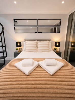 Ein modernes Hotelzimmer mit einem großen Bett, zwei Nachttischen mit Lampen, einem Spiegel über dem Bett und einer Wendeltreppe links.