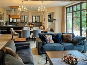 Un moderno salón con sofá, sillas y una mesa de centro. Cuenta con una zona de cocina con mesa y sillas. La habitación tiene ventanas amplias y una araña.
