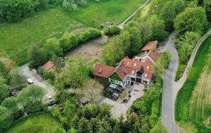 Een landhuis met een rood dak, omgeven door groen en een weg.