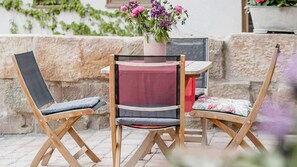 Una mesa de madera y dos sillas con cojines, un florero con flores y un muro de piedra al fondo.