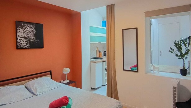 A hotel room with a bed, a bedside table, a mirror, a window with a plant, and a kitchenette in the background. - Modern Studio in Downtown Roubaix With High Speed Internet and Workspace (Roubaix)