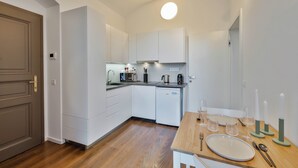 Cocina compacta con armarios blancos, lavavajillas integrado y una mesa de comedor de madera dispuesta con un plato, cubiertos y vasos.
