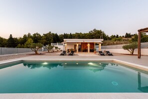 Un espace moderne aménagé au bord de la piscine, avec une piscine à eau claire, des chaises longues et un bâtiment doté d’un patio ouvert.