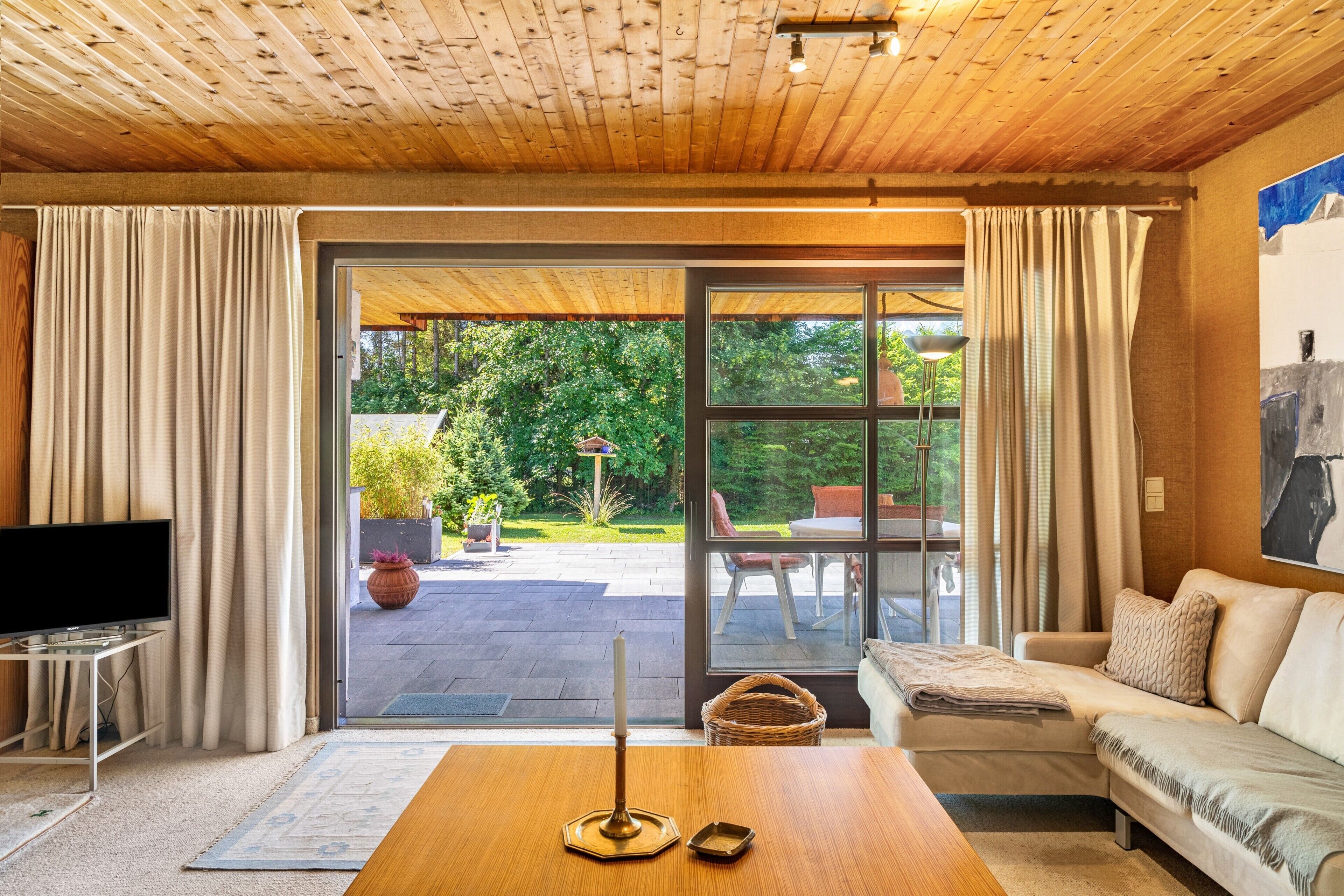 Un soggiorno con soffitto in legno, un divano, una TV su un mobile e vista su un giardino attraverso una porta in vetro.