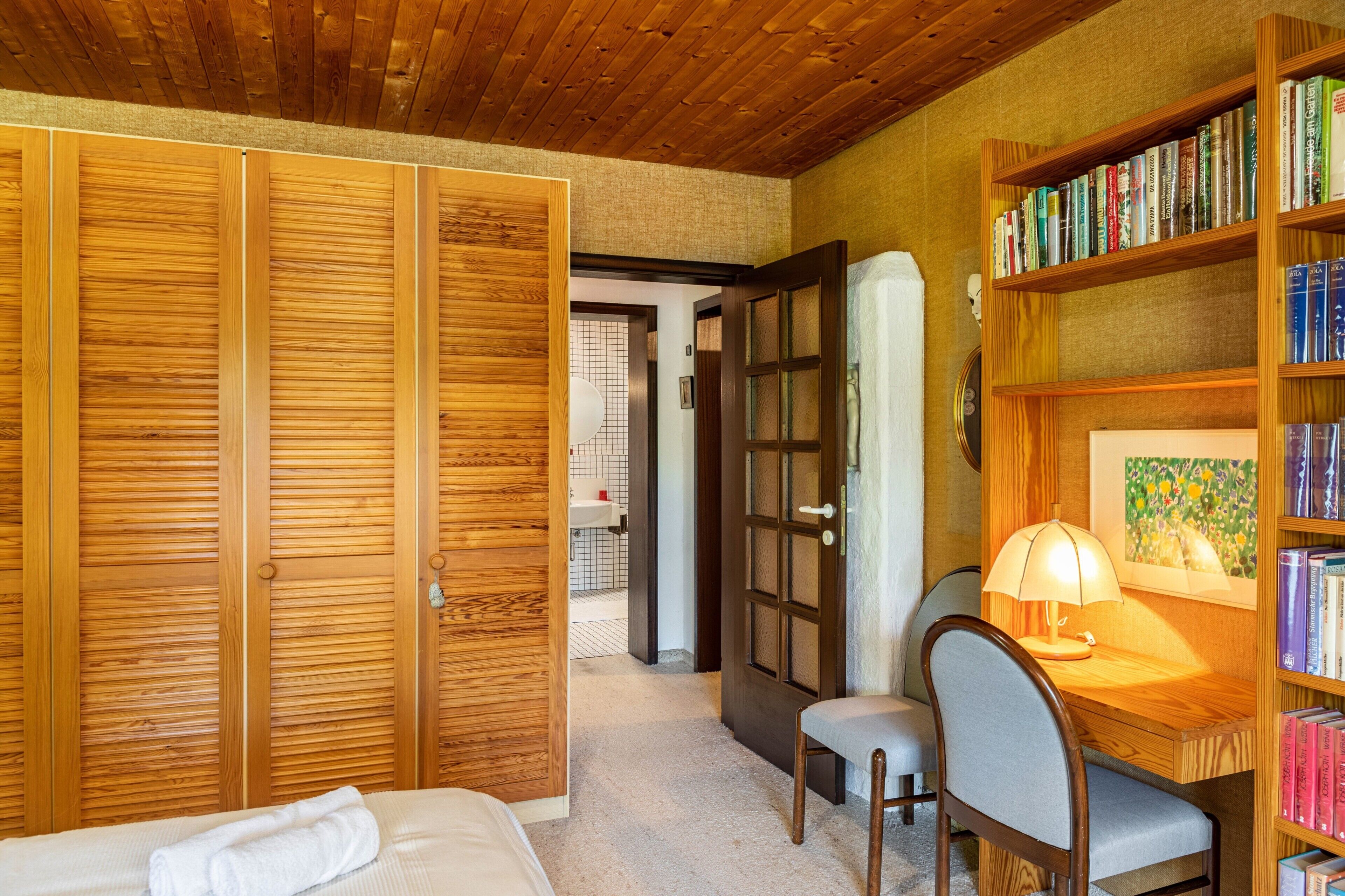 Una stanza con pannelli di legno, una libreria, una scrivania con una lampada, una sedia e un letto con un asciugamano arrotolato.