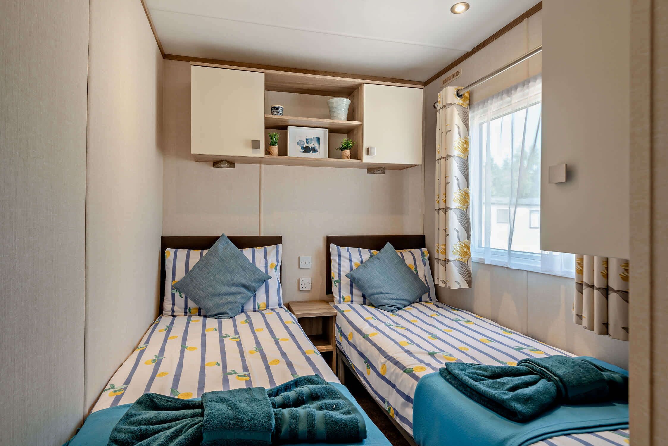 A compact hotel room with two single beds, a small bedside table, a built-in shelf with a plant, and a window with curtains.