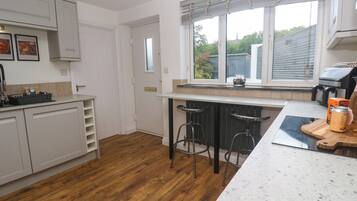 Cocina con alacenas blancas, piso de madera, ventana con persianas y encimera con una lata de café y una tabla de cortar.