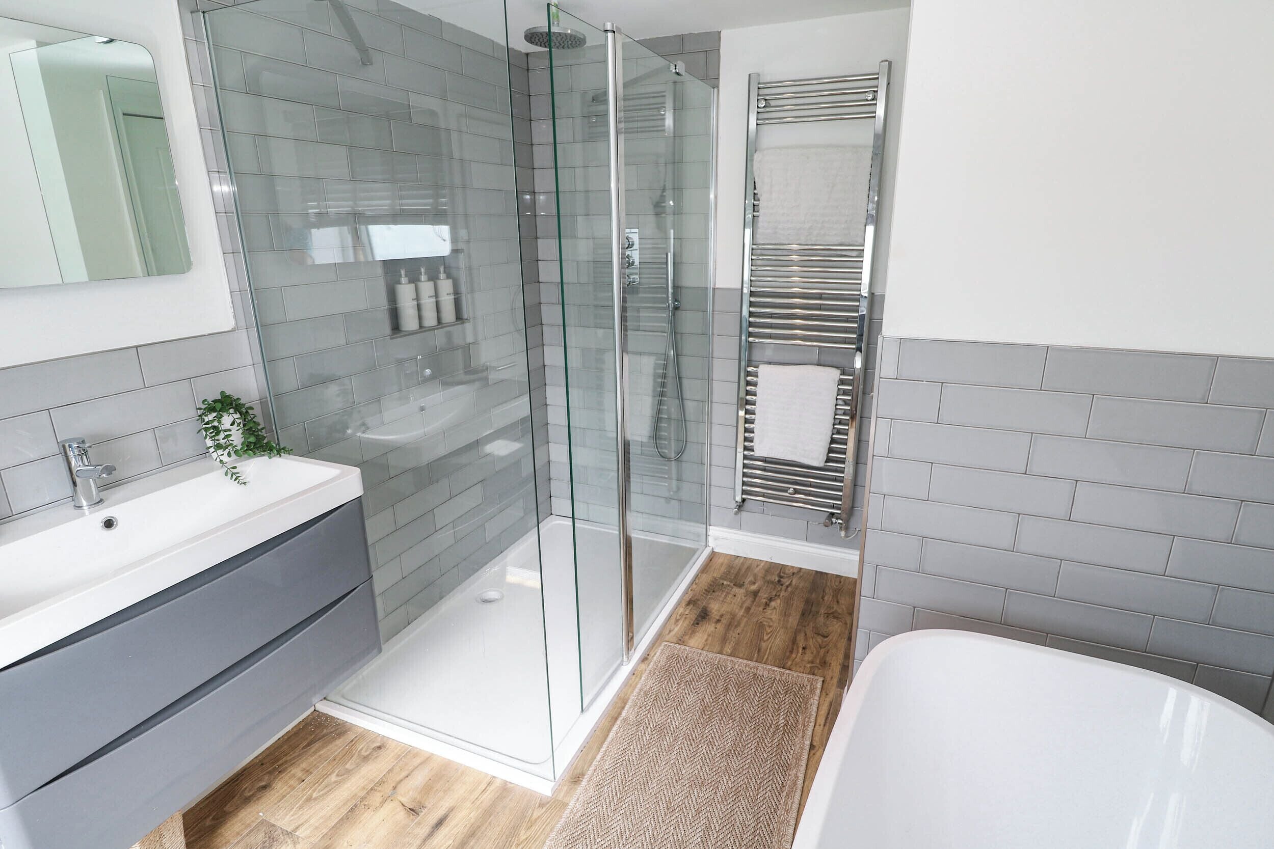 Un baño moderno con ducha envuelta en vidrio, una bañera blanca y un mueble de tocador con azulejos grises.