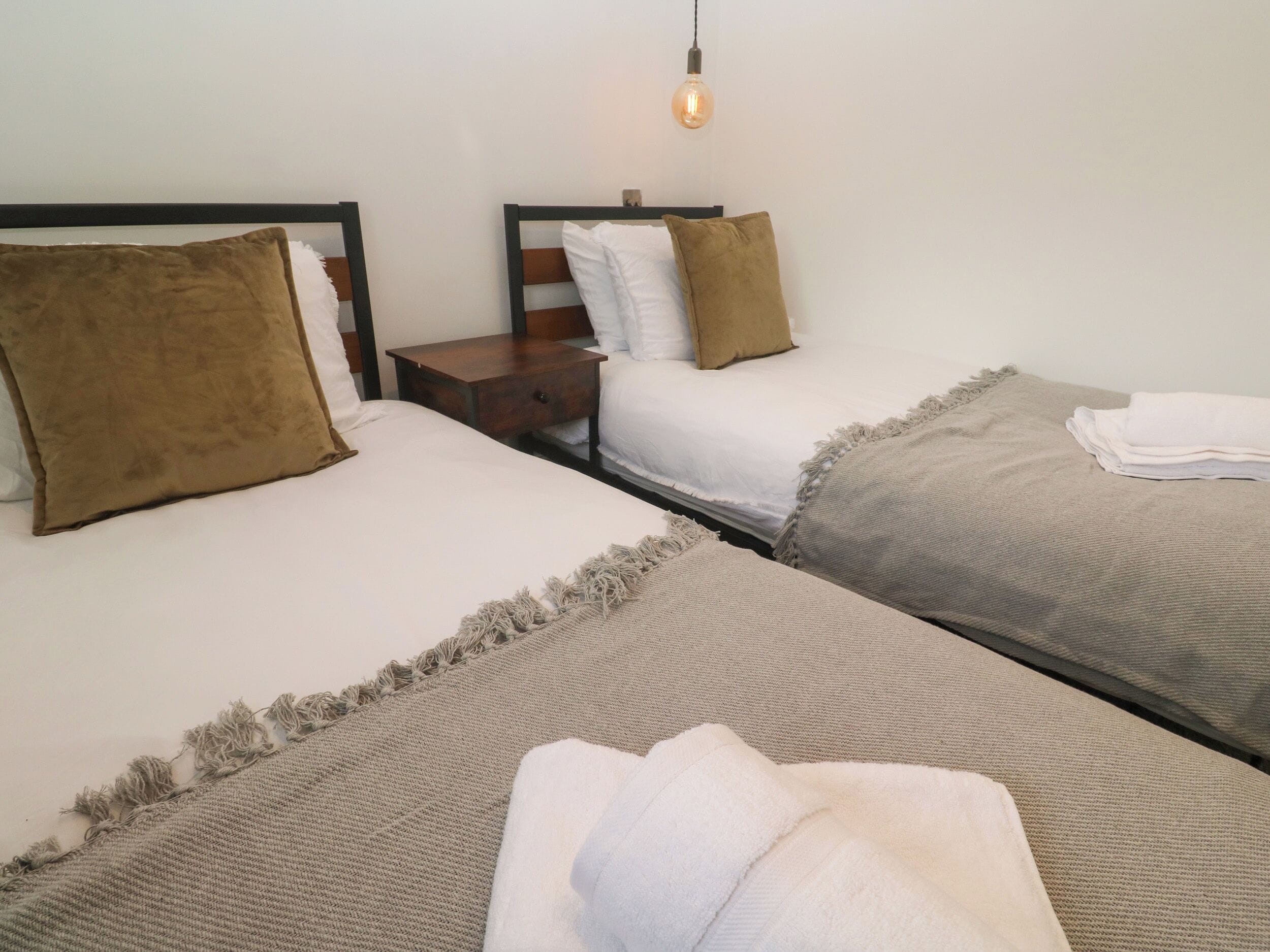 Una camera d'albergo con due letti, ognuno con un cuscino e una coperta. Tra i letti c'è un comodino con una lampada.