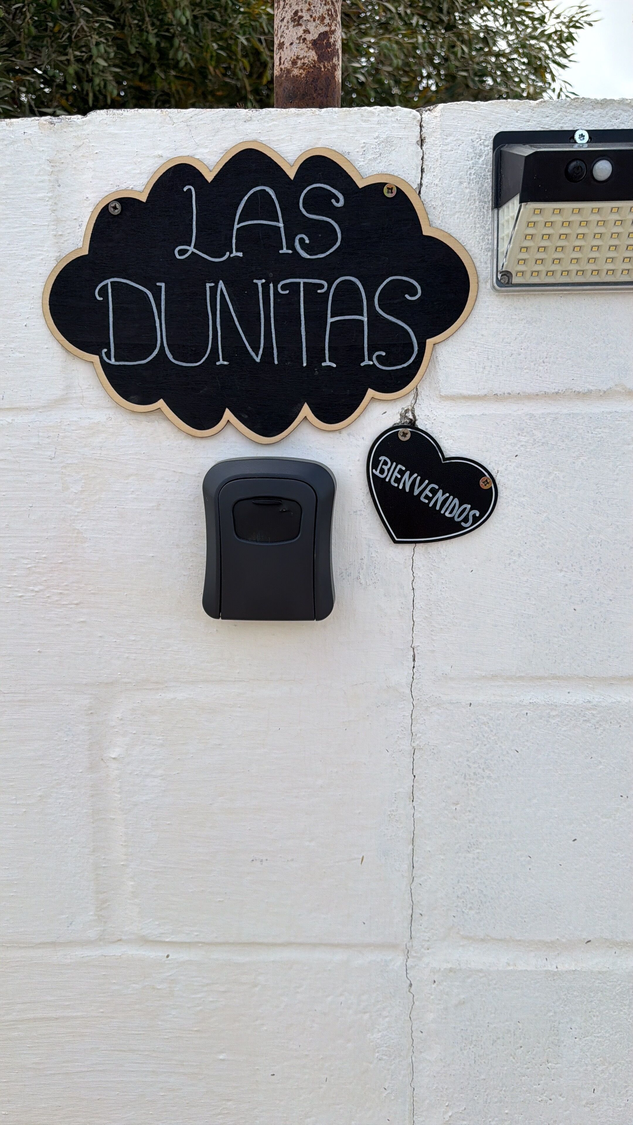 Un letrero en la pared con 'LAS DUNTAS' y la etiqueta 'BIENVENIDOS', un portero automático negro y una pared con textura.
