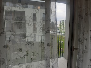 A view through a window with floral curtains, a balcony with a railing, and a building with windows in the background. - Beautiful apartment 20 minutes from downtown with parking (Warszawa)