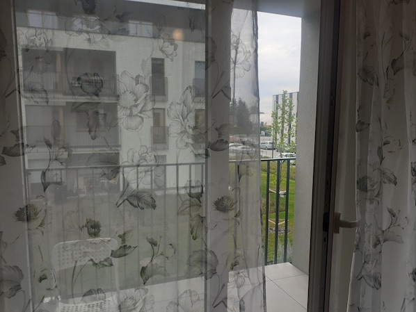 A view through a window with floral curtains, a balcony with a railing, and a building with windows in the background.