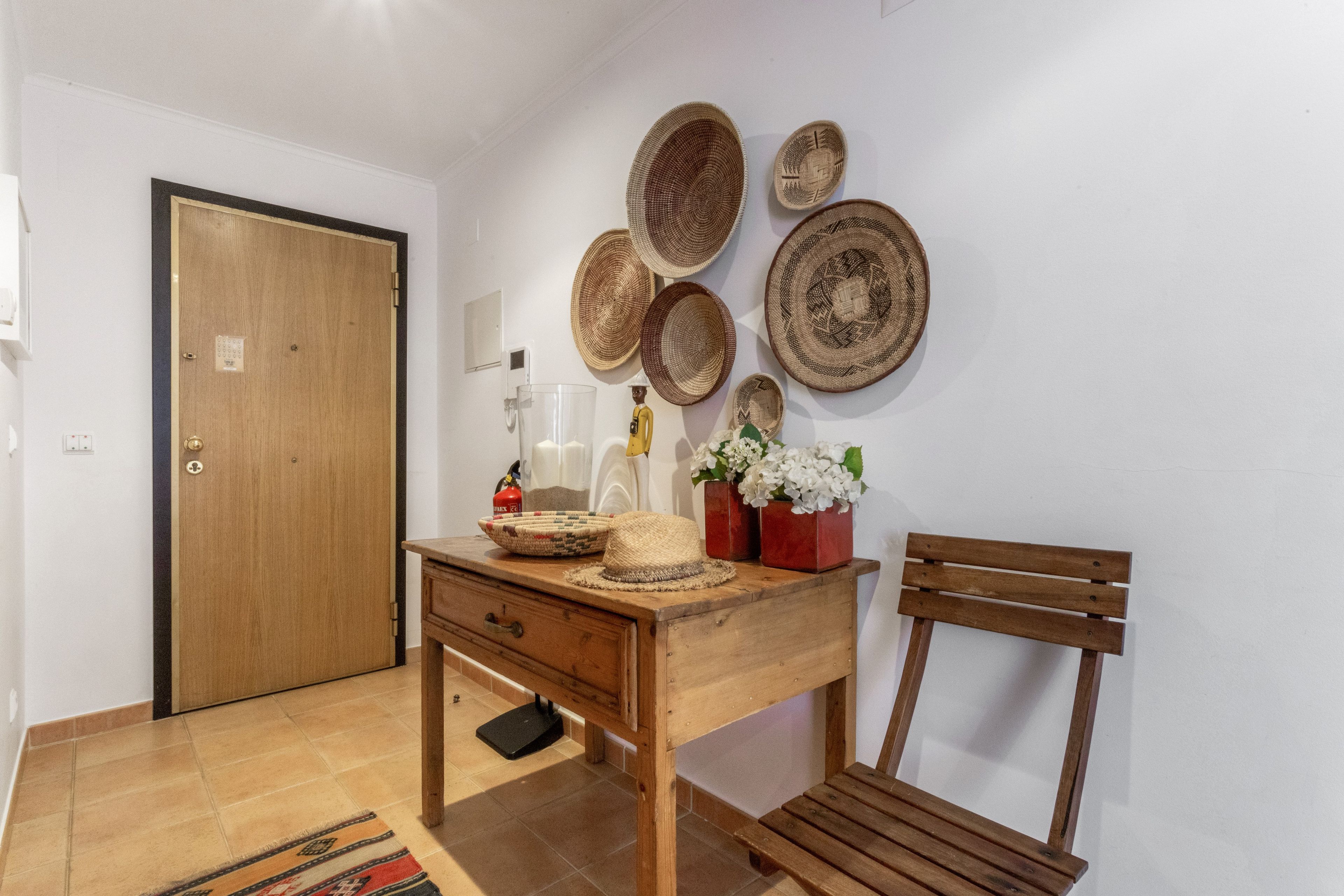 A room with a wooden door, a desk with a basket, a vase with flowers, and woven baskets on the wall.