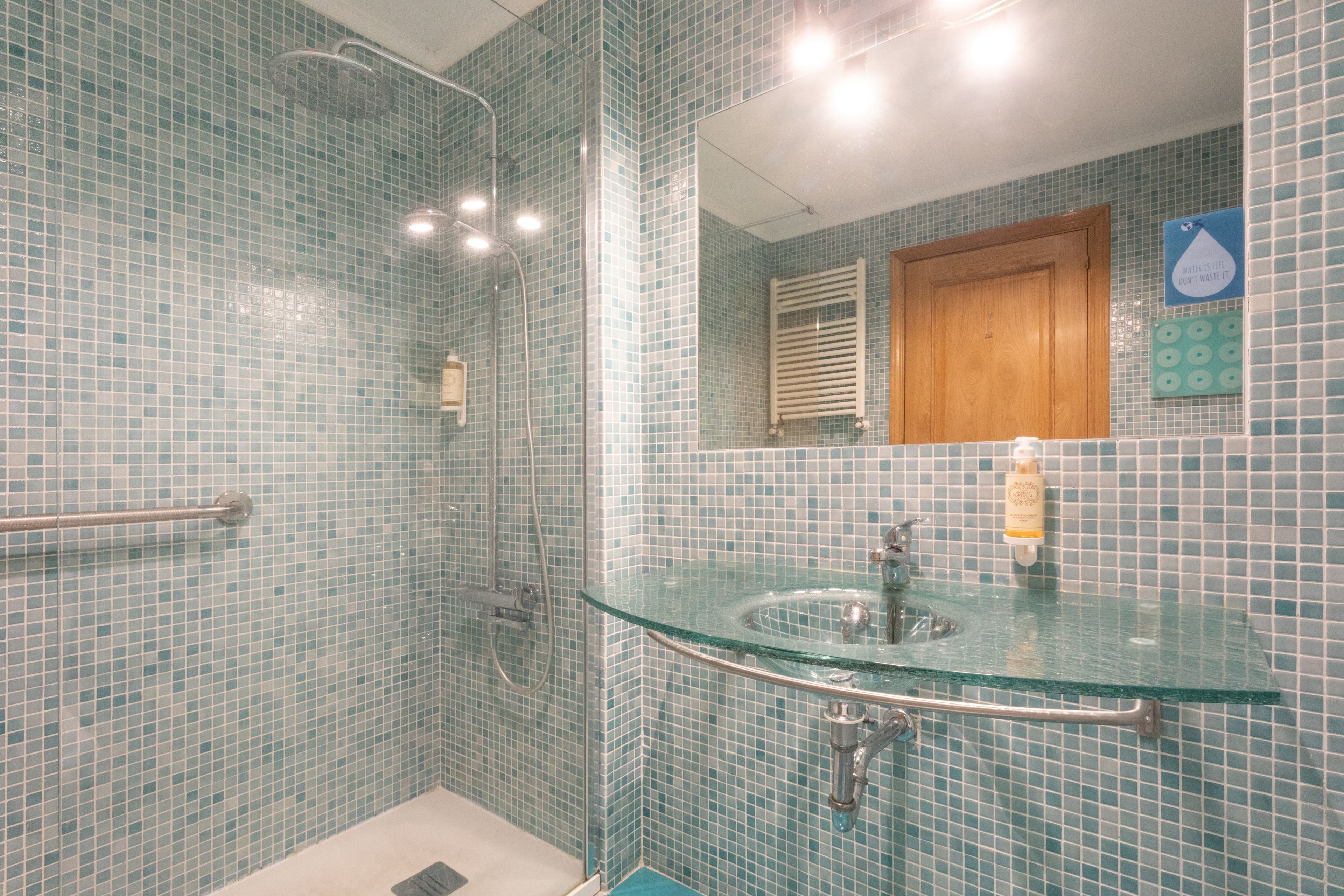 Un baño con lavamanos de vidrio, ducha con mampara de vidrio y puerta de madera.