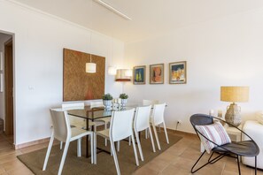 Ein moderner Essbereich mit einem Glastisch, weißen Stühlen und einem Teppich. An der Wand hängen drei gerahmte Gemälde und auf einem Beistelltisch steht eine Lampe.