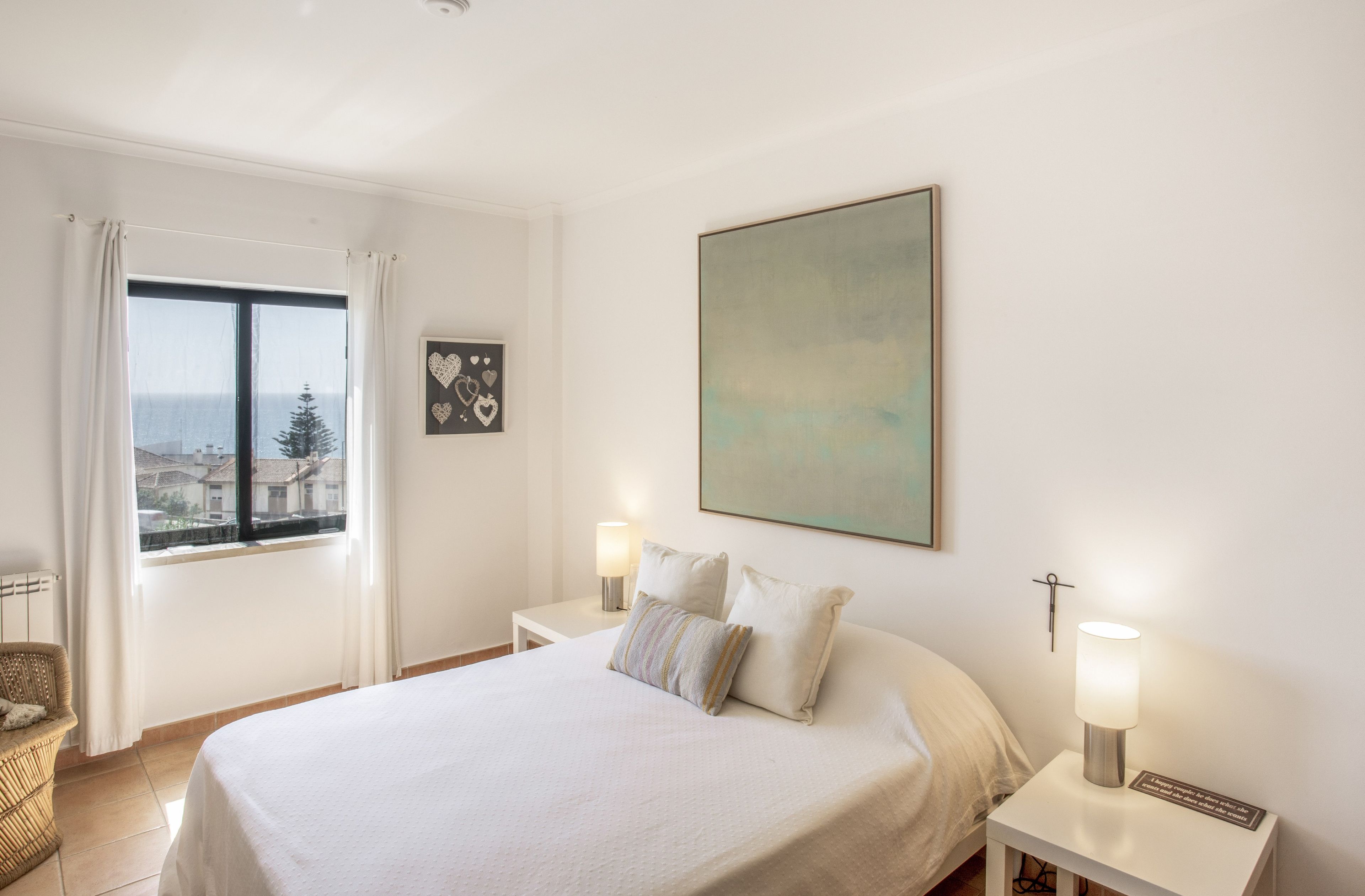 Una habitación de hotel con una cama, dos mesitas de noche con lámparas, un ventanal con vistas a edificios y un cuadro en la pared.