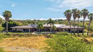 A two-story house with a patio, surrounded by palm trees and a hilly landscape. - Hula Hula Ranch - 7 Bedrooms, Pool, Ocean View! (Kamuela)