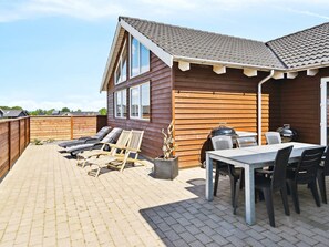 Ein Holzhaus mit einer Terrassenanlage, inklusive Tisch und Stühlen, Grill und Liegestühlen.