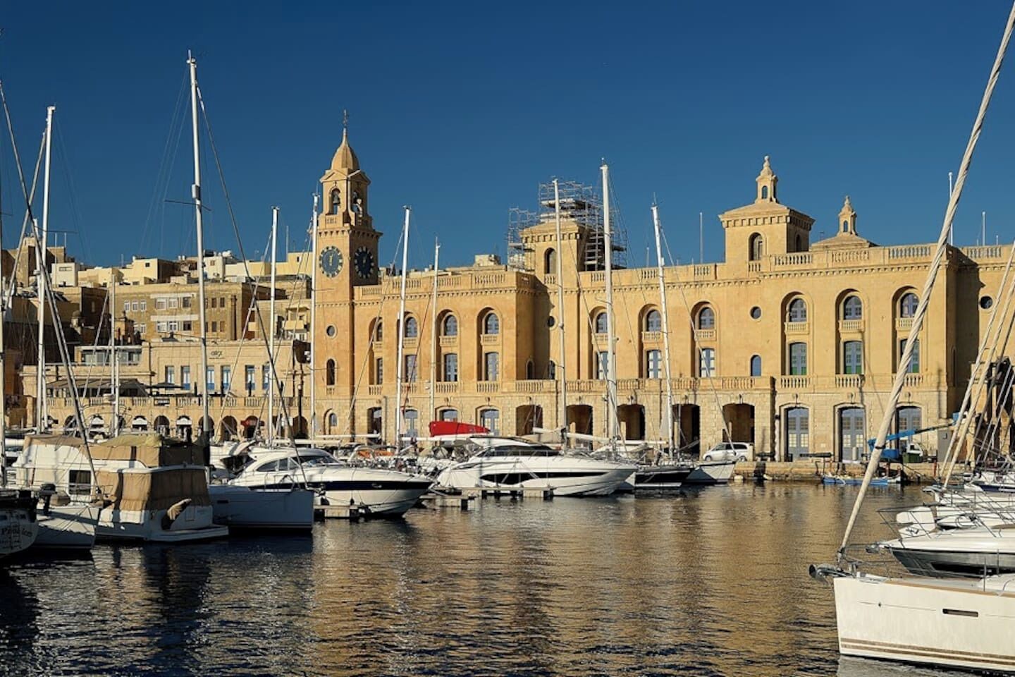 Un puerto deportivo con varias embarcaciones amarradas, una imponente torre del reloj y un edificio grande con ventanas de arco.