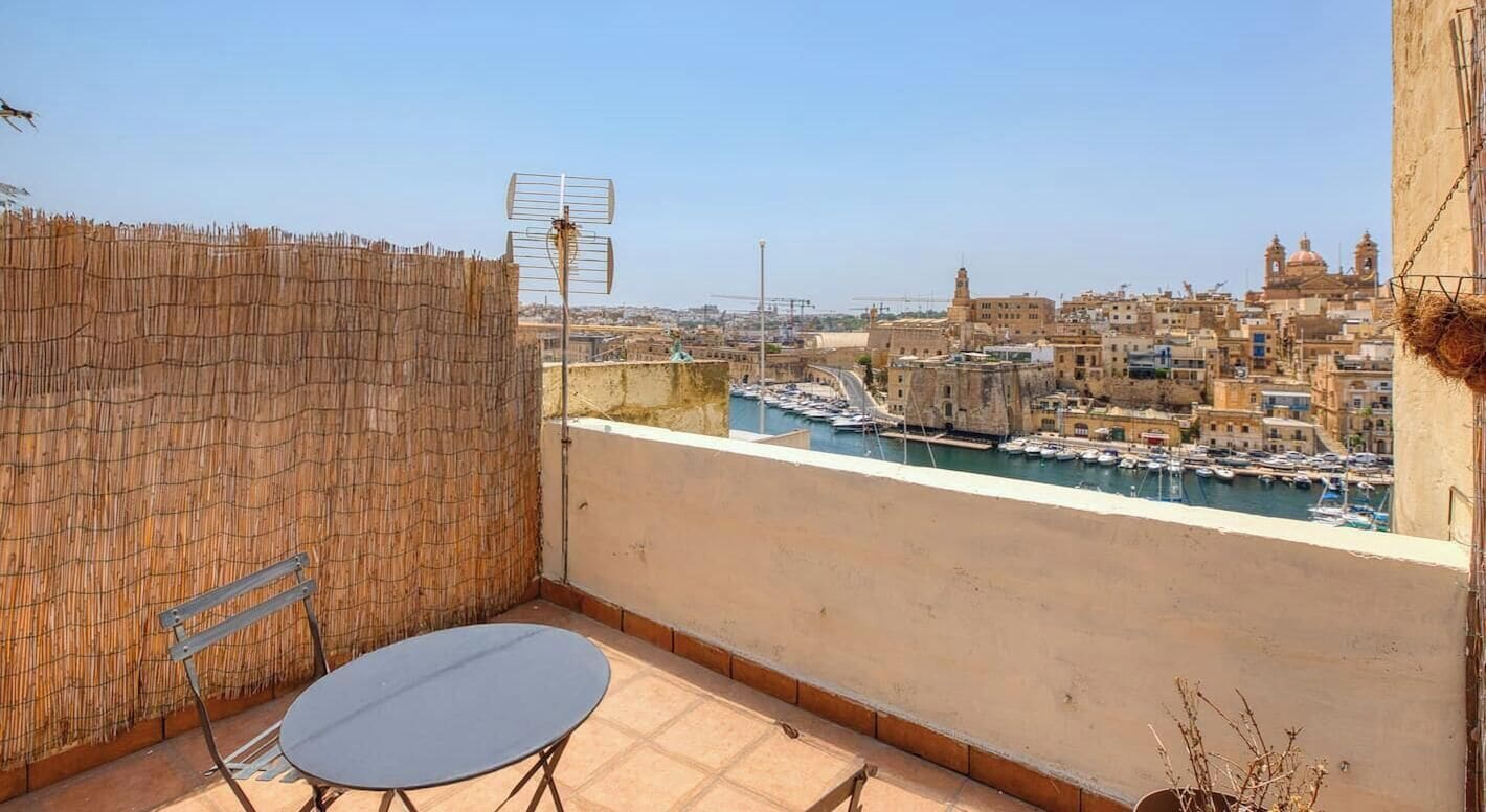 A balcony with a table and chairs, overlooking a marina with boats and a cityscape.