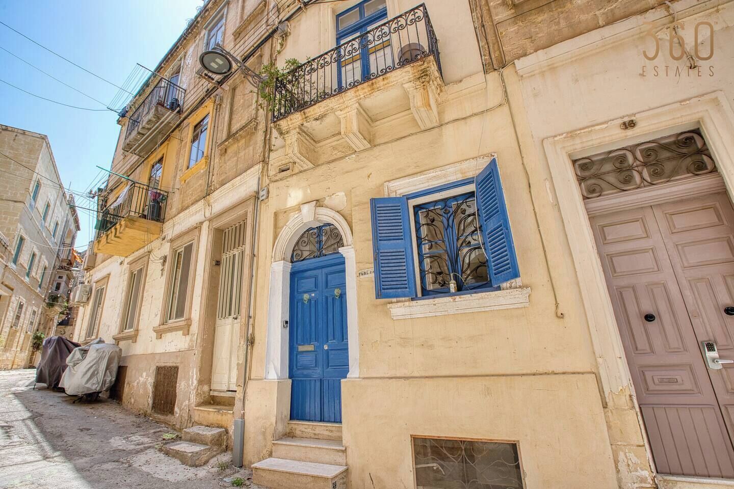 A narrow street with old buildings, a blue door, and a balcony with a wrought iron railing.