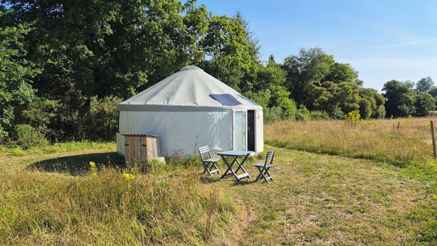 Ein kleines Zelt für eine Person, mit Tür und Fenster, umgeben von einer grünen Wiese mit einigen Stühlen und einem Tisch.