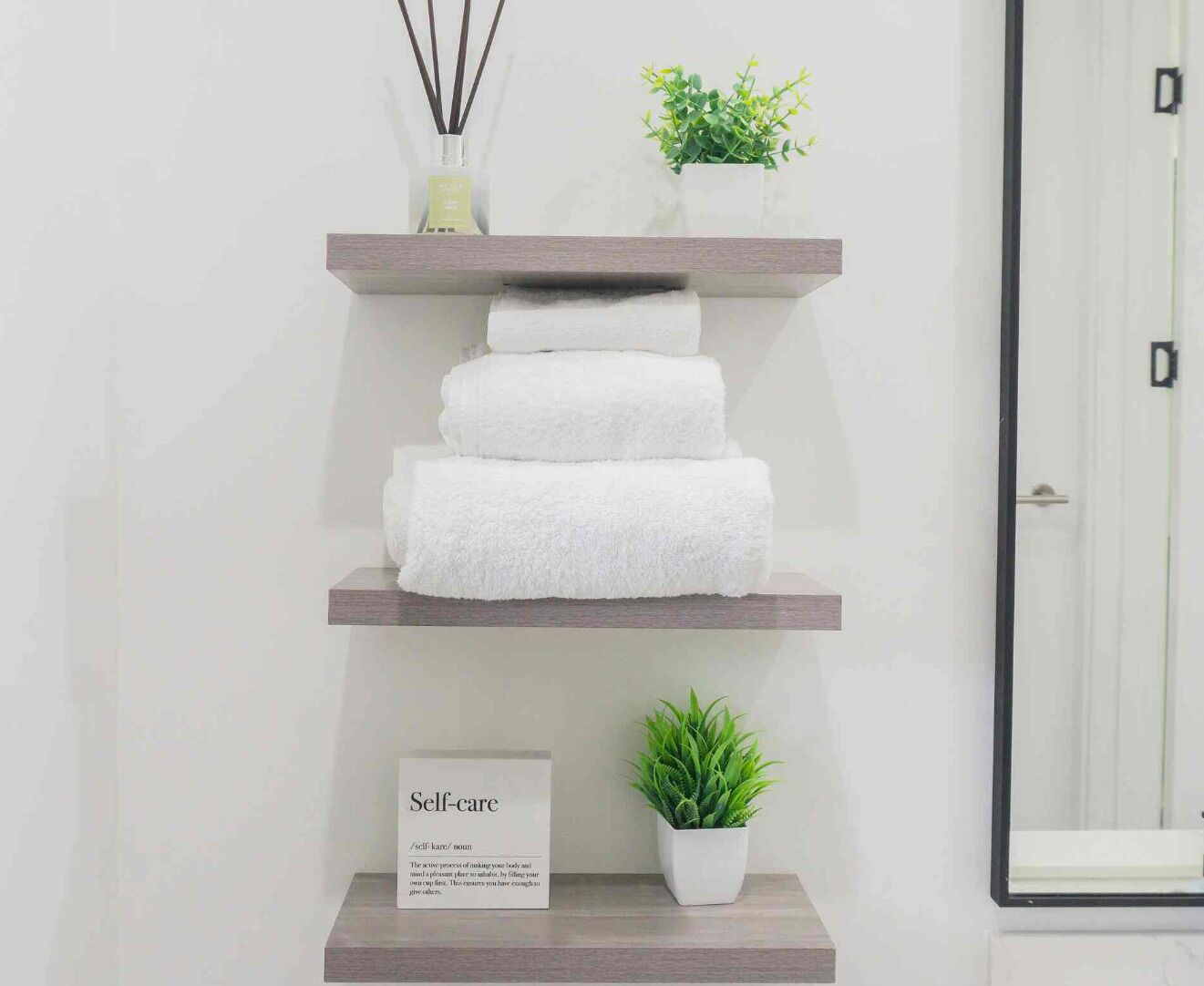 Un baño con un estante de madera que sostiene toallas blancas, una pequeña planta en maceta y un cartel con la palabra 'Self-care' y una definición.