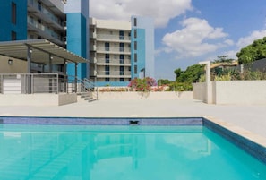 Una piscina con superficie de agua azul cristalina, rodeada por una zona exterior cubierta con una escalera que conduce a un edificio con varios balcones.