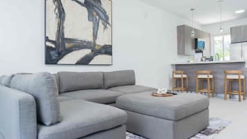 Ein modernes Wohnzimmer mit Ecksofa und Hocker, einem großen abstrakten Gemälde und einer Küchenzeile mit Barhocker.