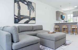 Una sala de estar moderna con un sofá seccional y un reposapiés, un cuadro abstracto grande y una zona de cocina con taburetes de bar.