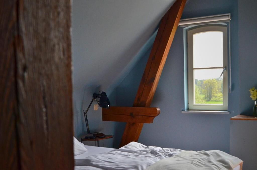 Un dormitorio con techo inclinado, ventana con vistas a zonas verdes, mesita de noche con lámpara y cama con ropa blanca.