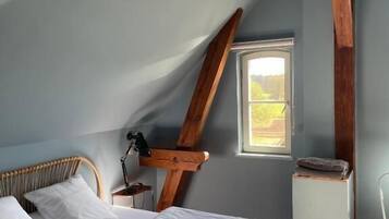 Un dormitorio con techo inclinado, una cama con ropa de cama blanca, una mesita de noche con lámpara, un ventanal y un radiador.