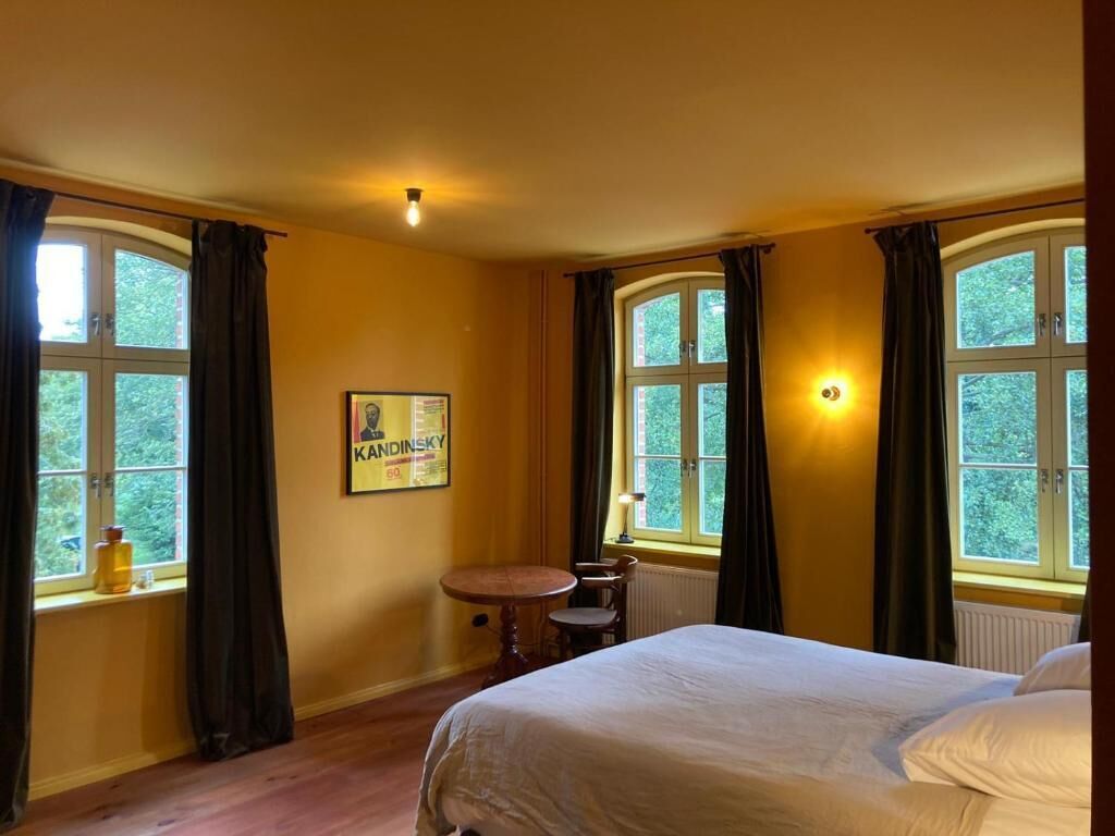 Une chambre d’hôtel avec un lit, une petite table, deux fenêtres avec des rideaux, un radiateur et un luminaire fixé au mur.