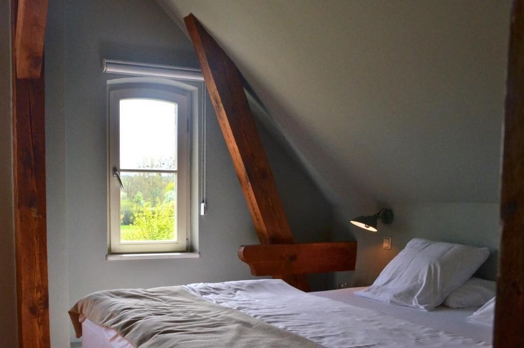 A bedroom with a sloped ceiling, a window with a view of greenery, a bed with white linens, and a wall-mounted reading light.