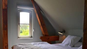 A bedroom with a sloped ceiling, a window with a view of greenery, a bed with white linens, and a wall-mounted reading light.