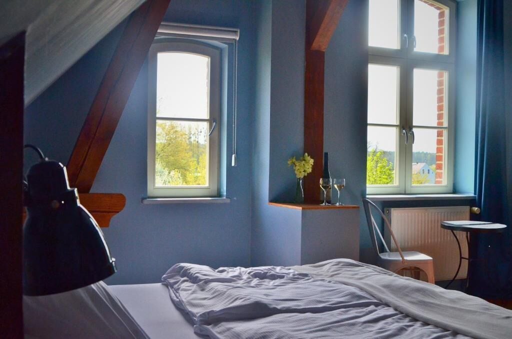 Ein Schlafzimmer mit einem Bett, zwei Fenstern, einem Sessel, einem kleinen Tisch und einer Heizung.