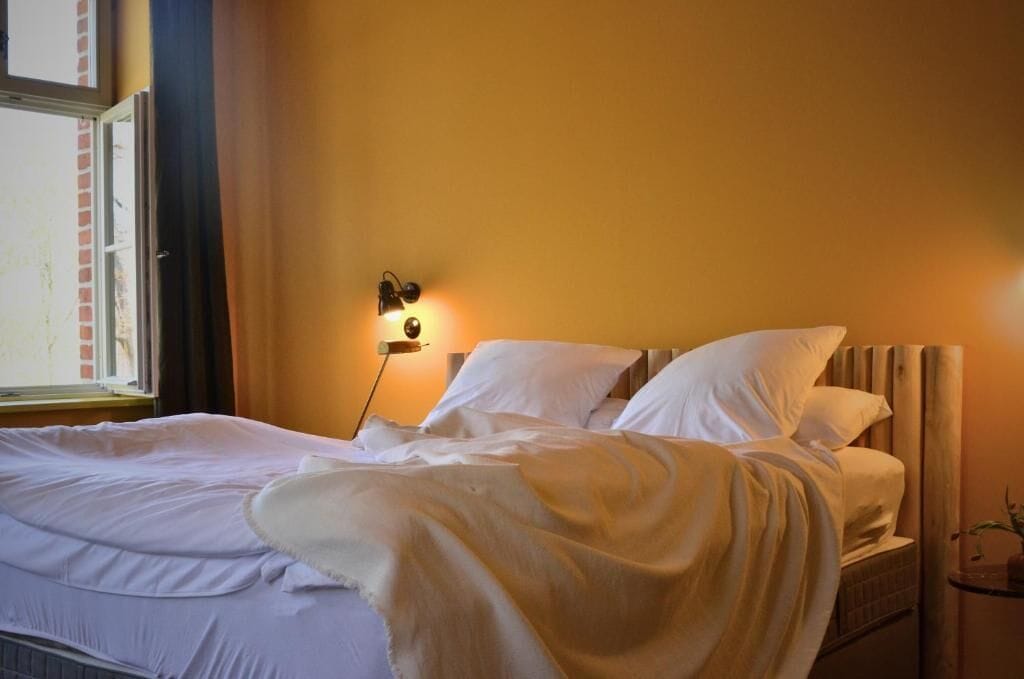 Ein ordentlich bezogenes Bett mit weißen Laken in einem Zimmer mit Fenster und Wandlampe.
